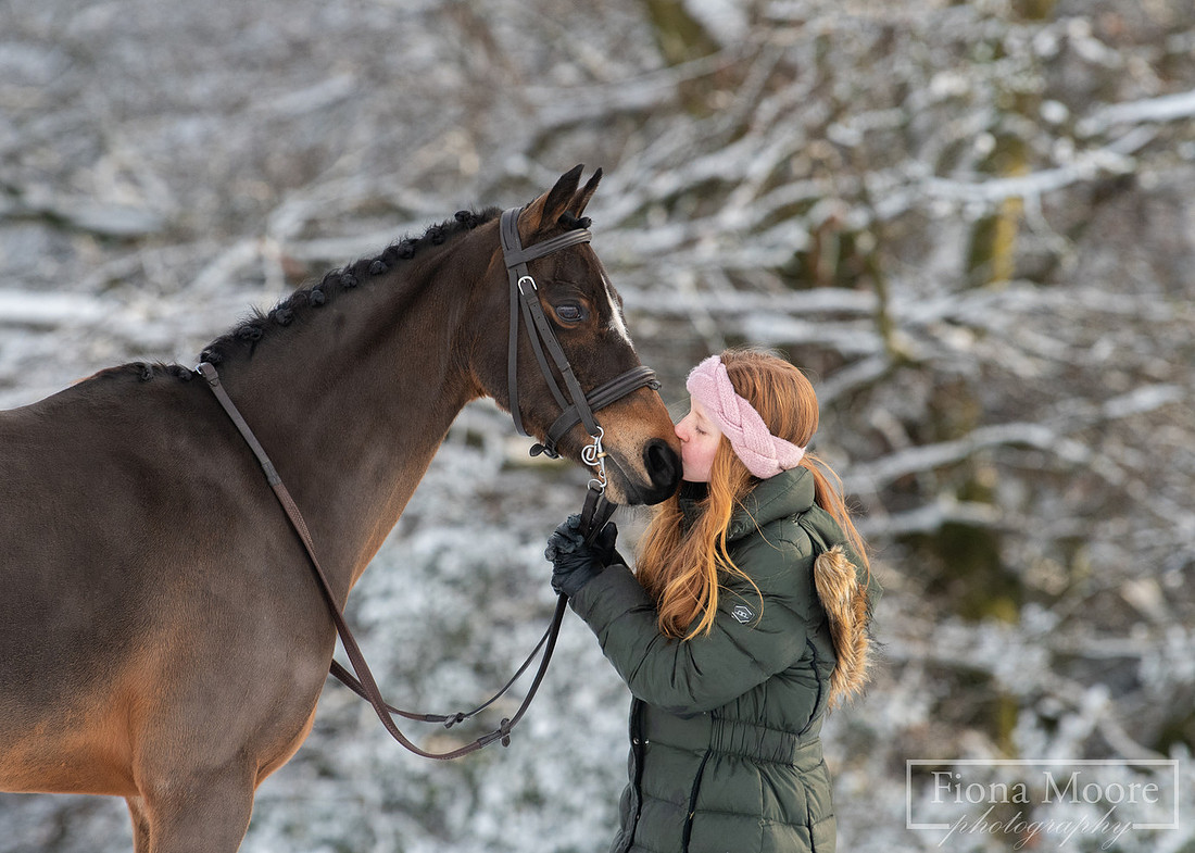 Portait of a young girl kissing her bay pony's nose in a winter landscape. Photograph taken by West Midlands Based Specialist Equine Photographer Fiona Moore Photography